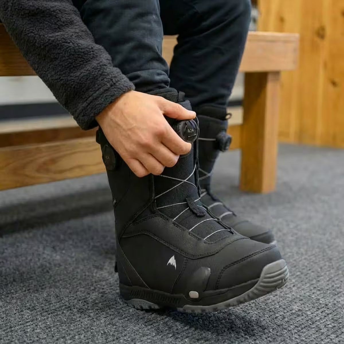 Person wearing black winter boots with a visible brand logo, sitting on a wooden bench.