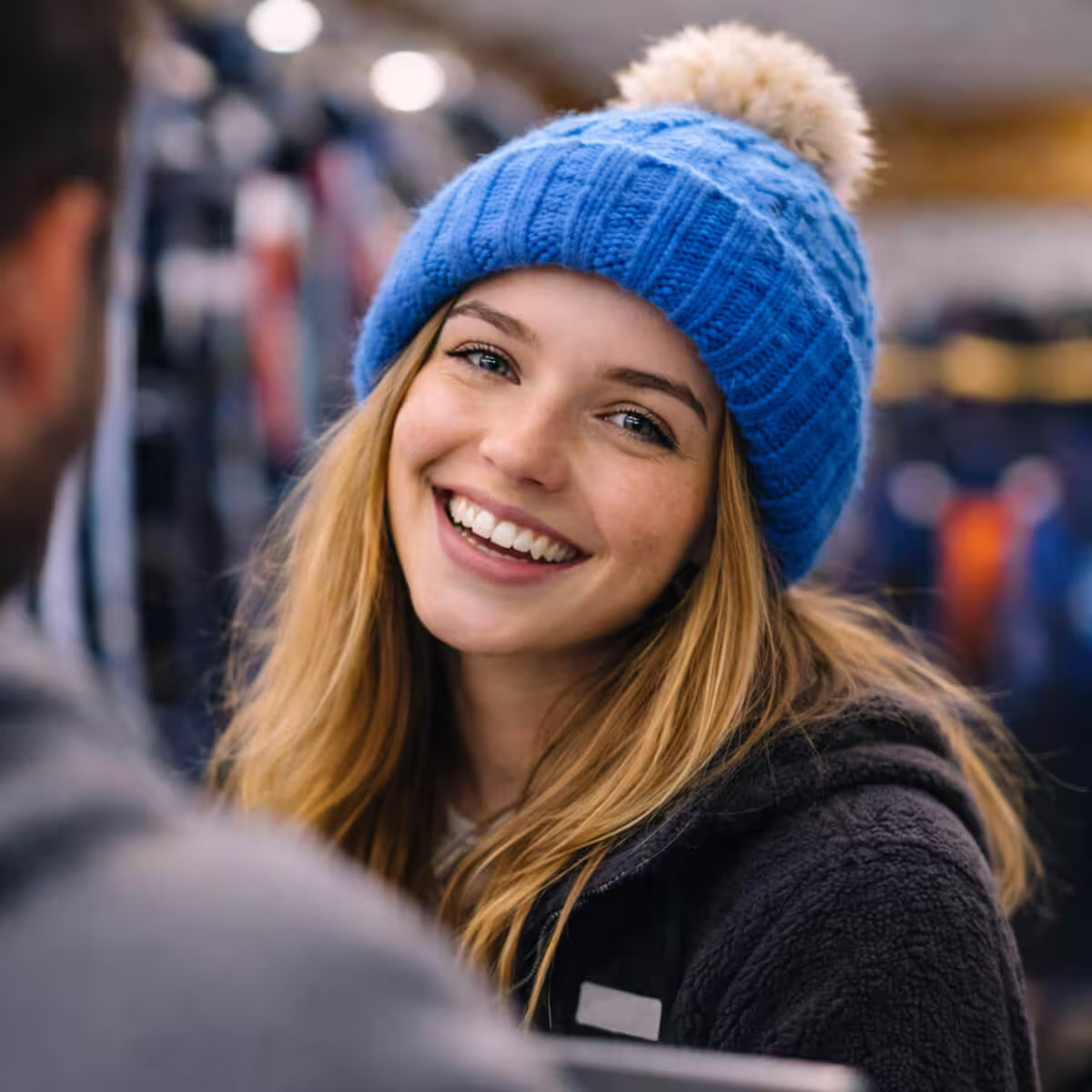 Woman wearing a blue knit hat with a pom-pom, smiling in an indoor setting.
