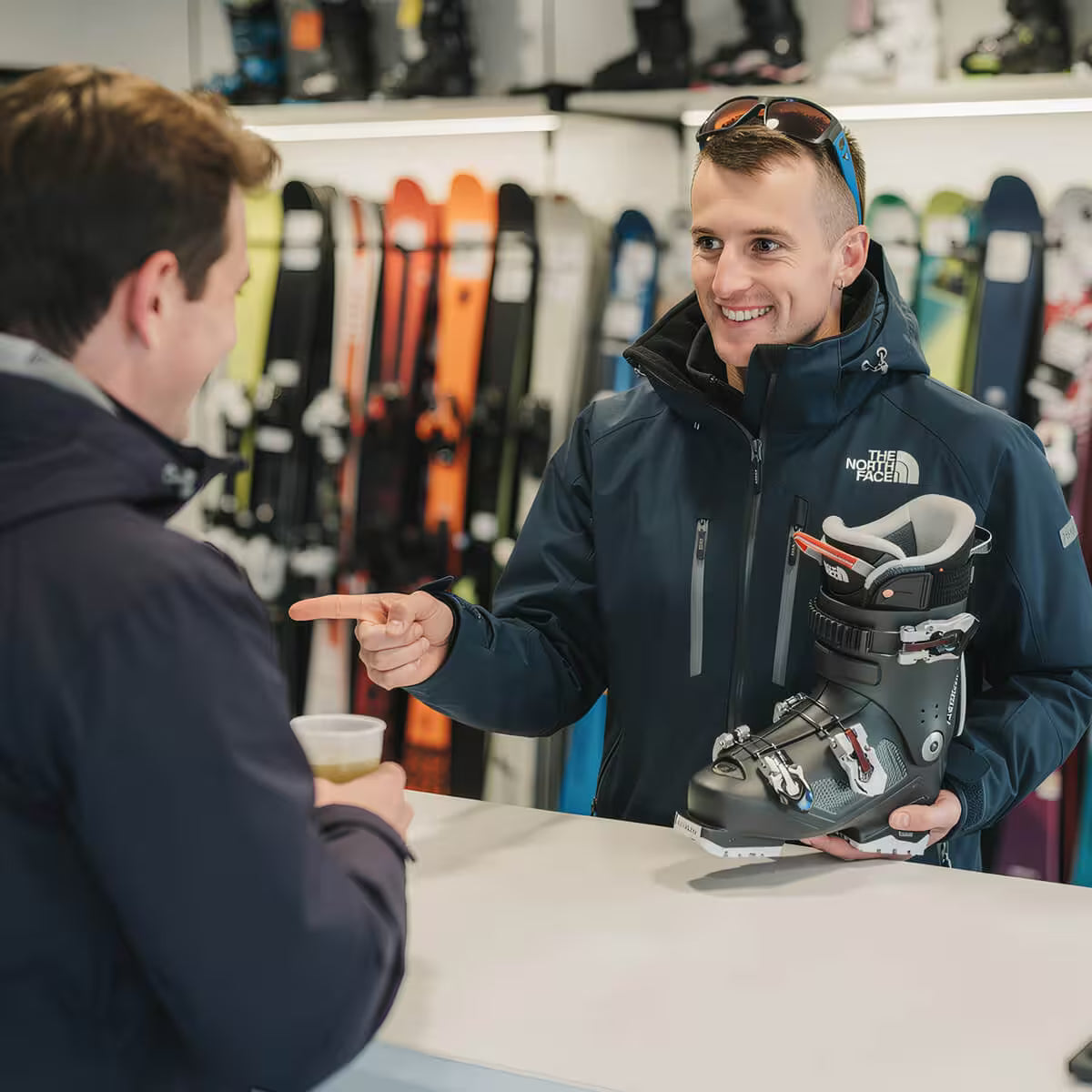 Person holding a ski boot and pointing at another person in a store with skis in the background