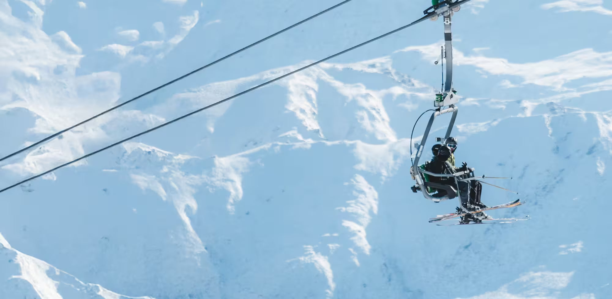 Two skiers on a chairlift against a snowy mountain backdrop