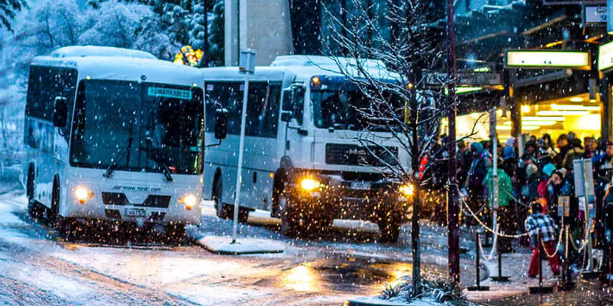 Two Queenstown ski buses in a snowy urban setting with people waiting at a bus stop.