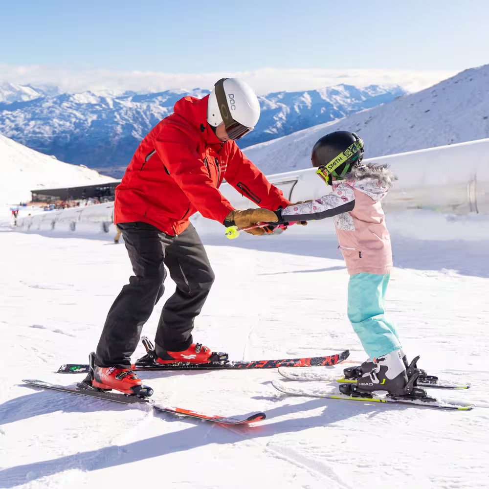 Person teaching a child skiing on a snowy mountain with skis and helmets.