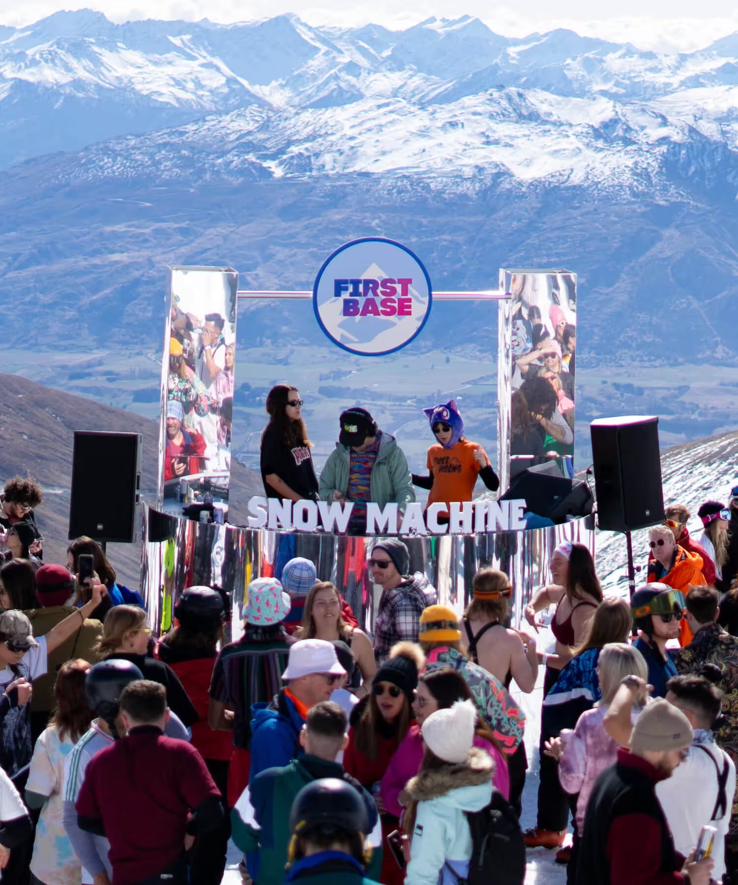 Crowd at a ski resort event with 'First Base' and 'Snow Machine' branding, mountains in the background.