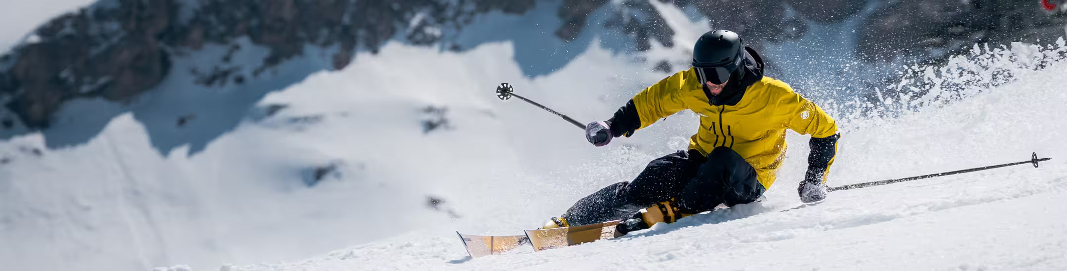 Skier in a yellow jacket skiing down a snowy slope with mountains in the background