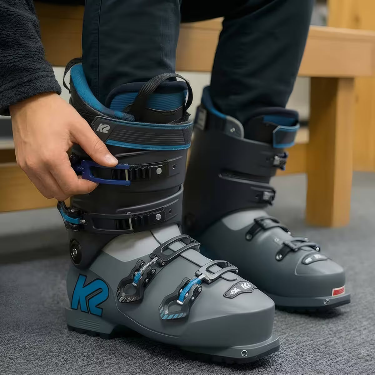 Person adjusting gray ski boots with blue accents on a carpeted floor.