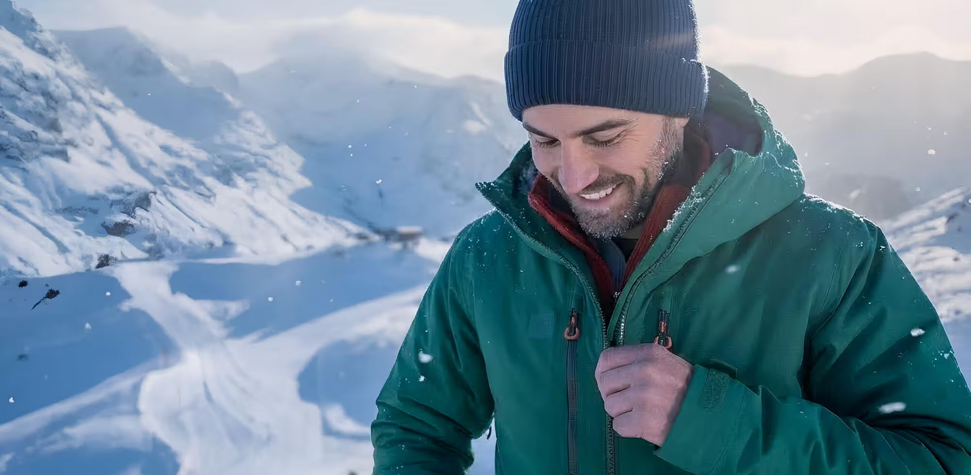Man in a green jacket and black beanie standing in a snowy landscape with mountains.
