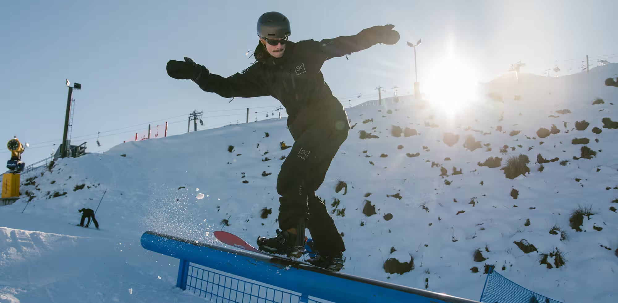 Person snowboarding on a rail at a snowy slope with a bright sun in the background