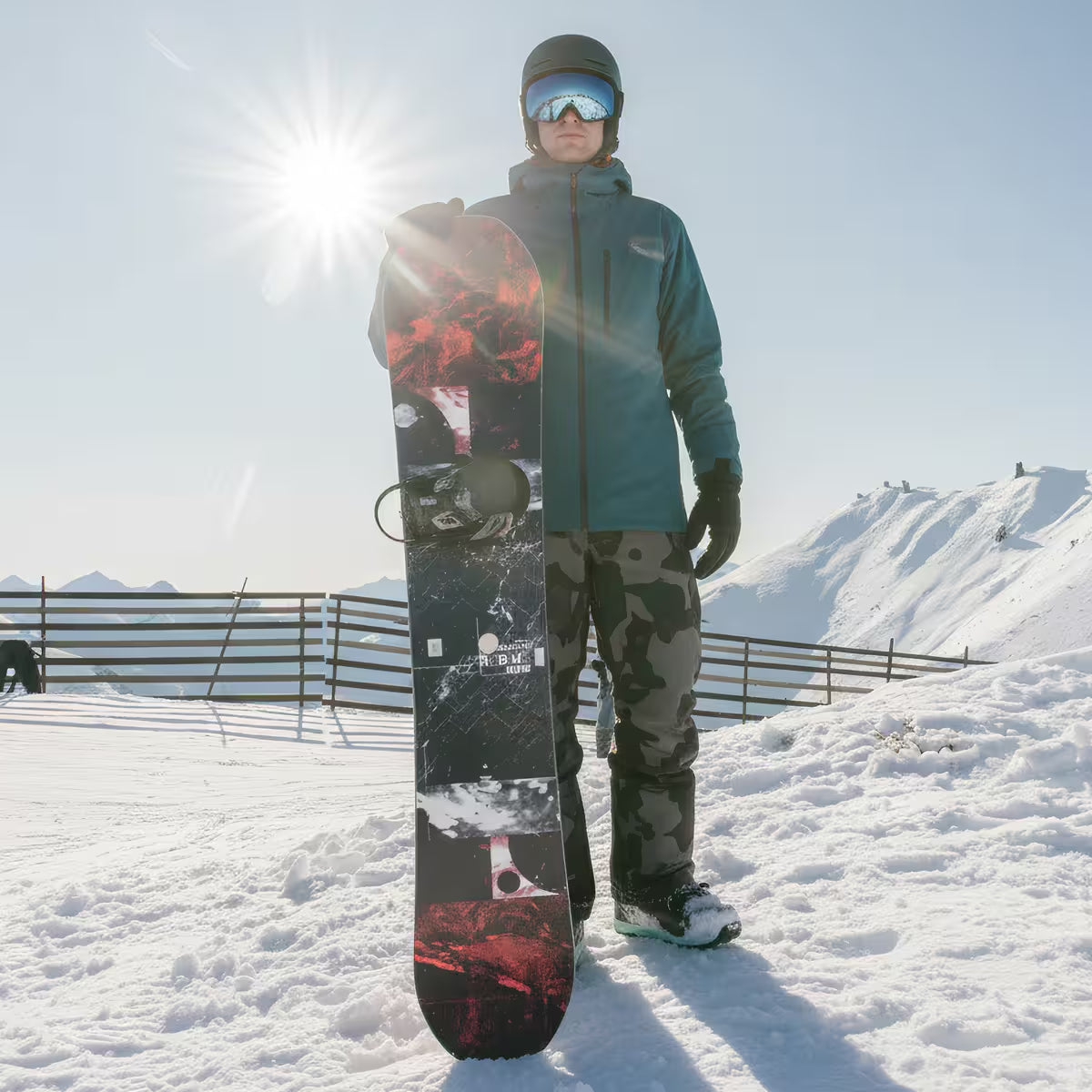 Person holding a snowboard in a snowy landscape with mountains in the background